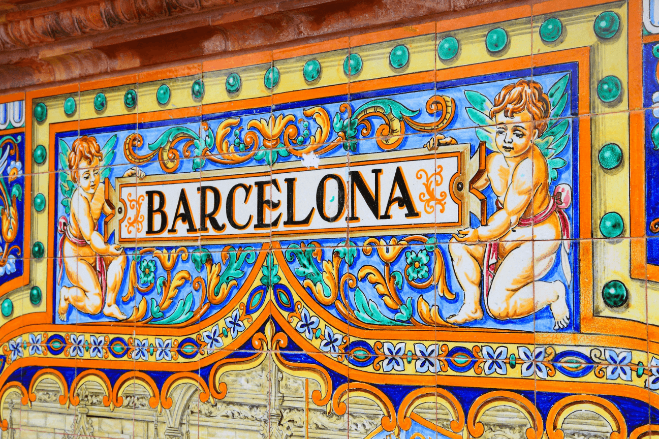 A sign saying Barcelona made of brightly coloured tiles. With floral decoration and a yellow border with blue circles, two putti hold up a tablet saying Barcelona