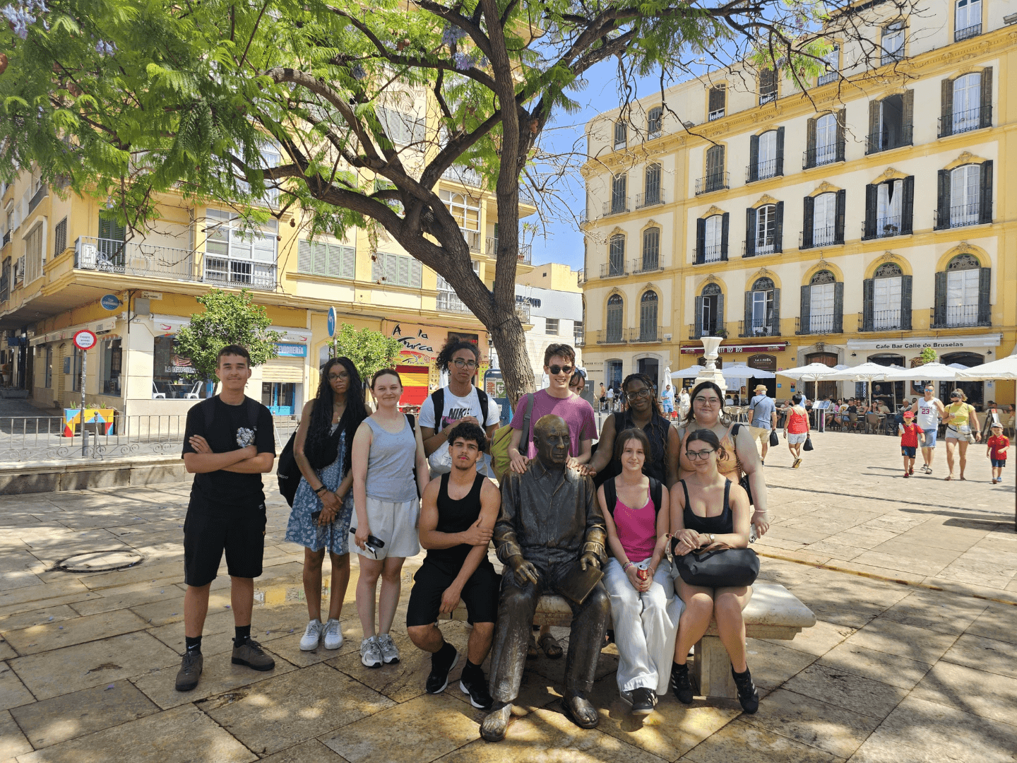 A group of young people with a statue of Pablo Picasso in a square in Malaga, shaded by a green-leaf tree.