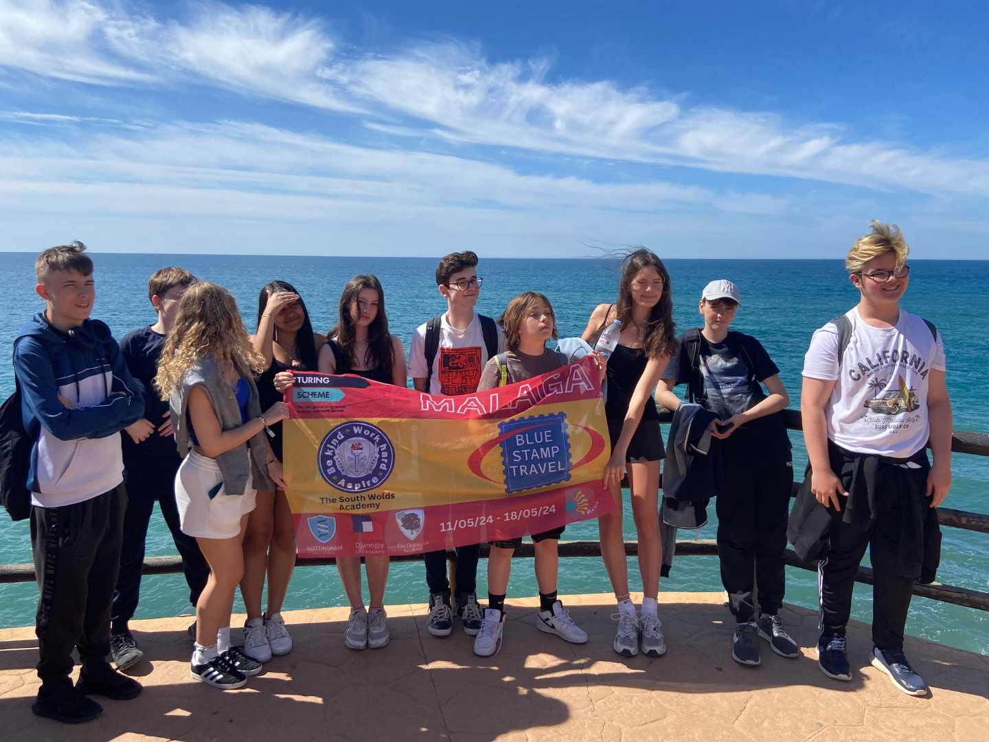 A group of young people on a school trip to Malaga hold up a Blue Stamp Travel banner with the sea behind them
