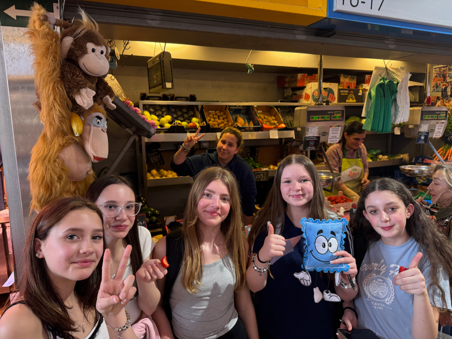 A group of students on a school trip to Malaga pose for a group photo in front of a market stall. One holds Stampie, the Blue Stamp Travel mascot.