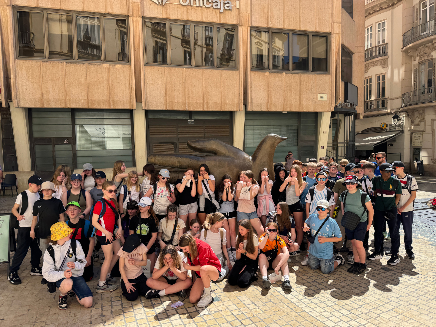 A large group of students on a school trip to Malaga pose for a group photo with a sculpture of a dove in the background.