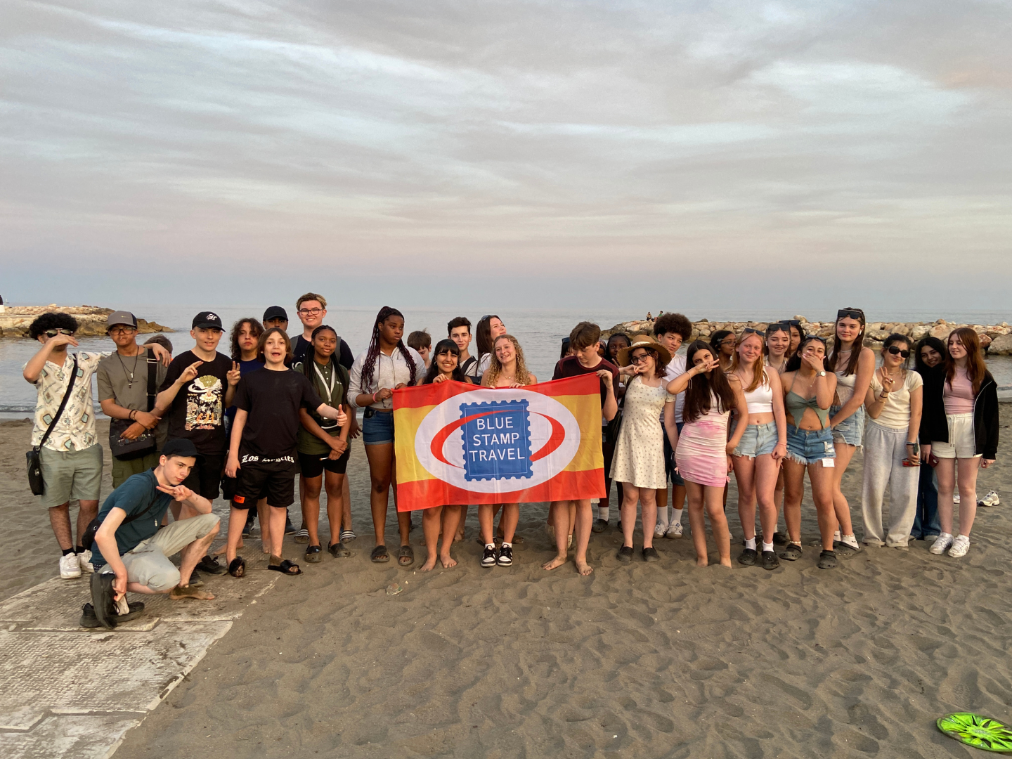 A group of students pose for a group photo on Malaga beach, holding a Blue Stamp Travel banner
