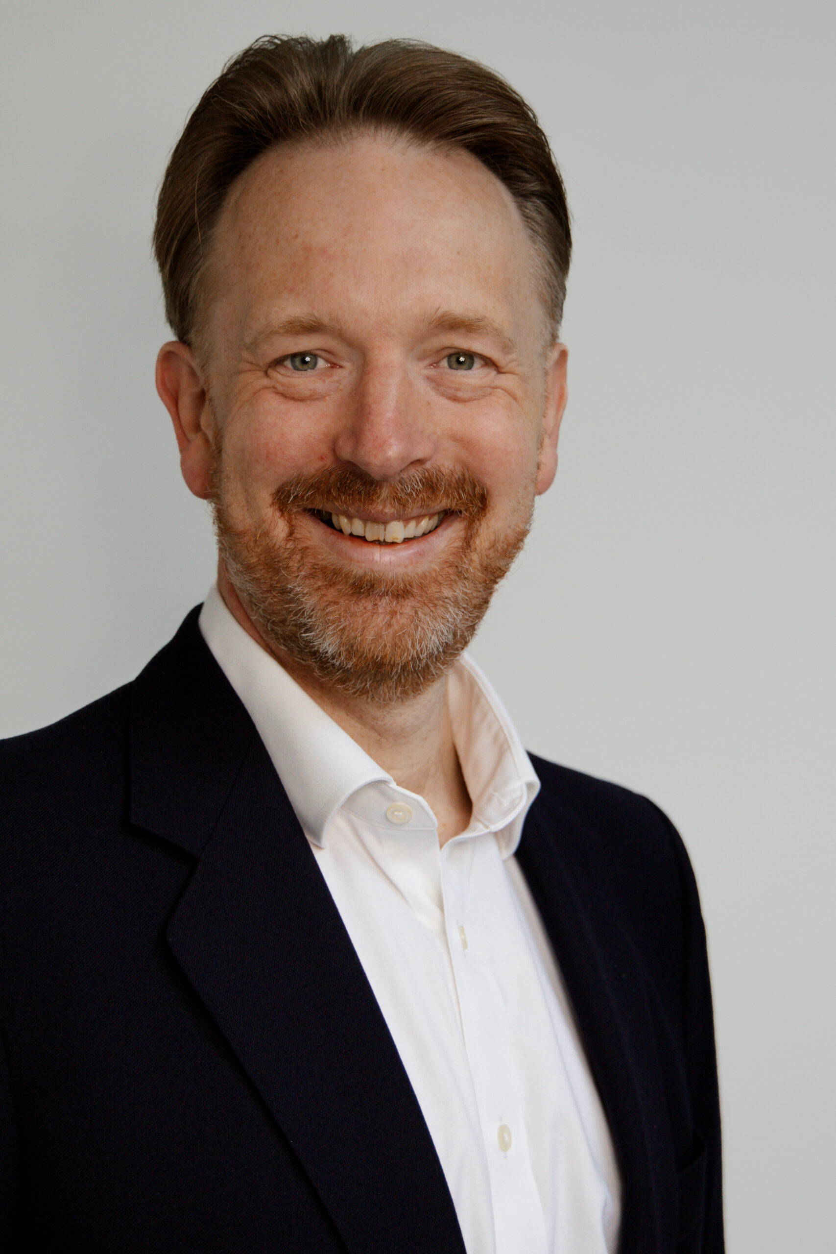 A man in his forties wearing a white shirt and dark blazer.